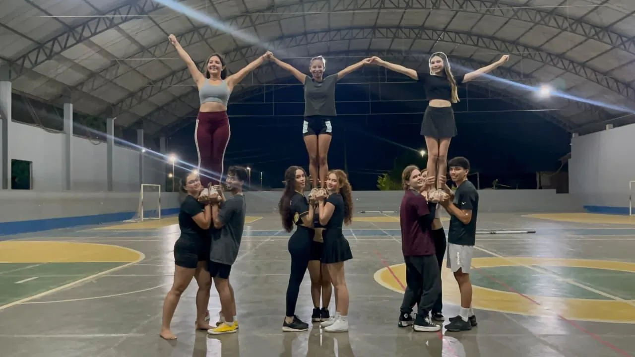 Três atletas da equipe no alto de uma pirâmide humana, numa quadra coberta à noite